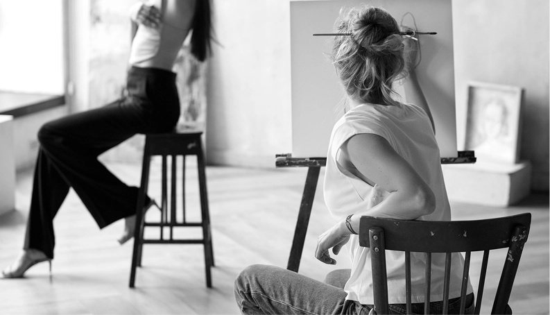 Two women seated on chairs, engaged in discussion, with an easel displaying artwork in front of them.